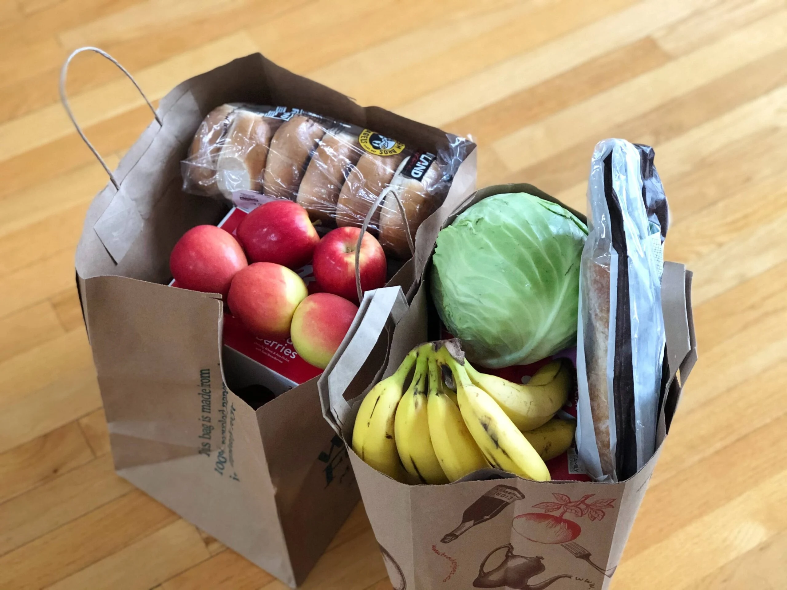 Two full grocery bags with apples, bananas, bagels and cabbage on top