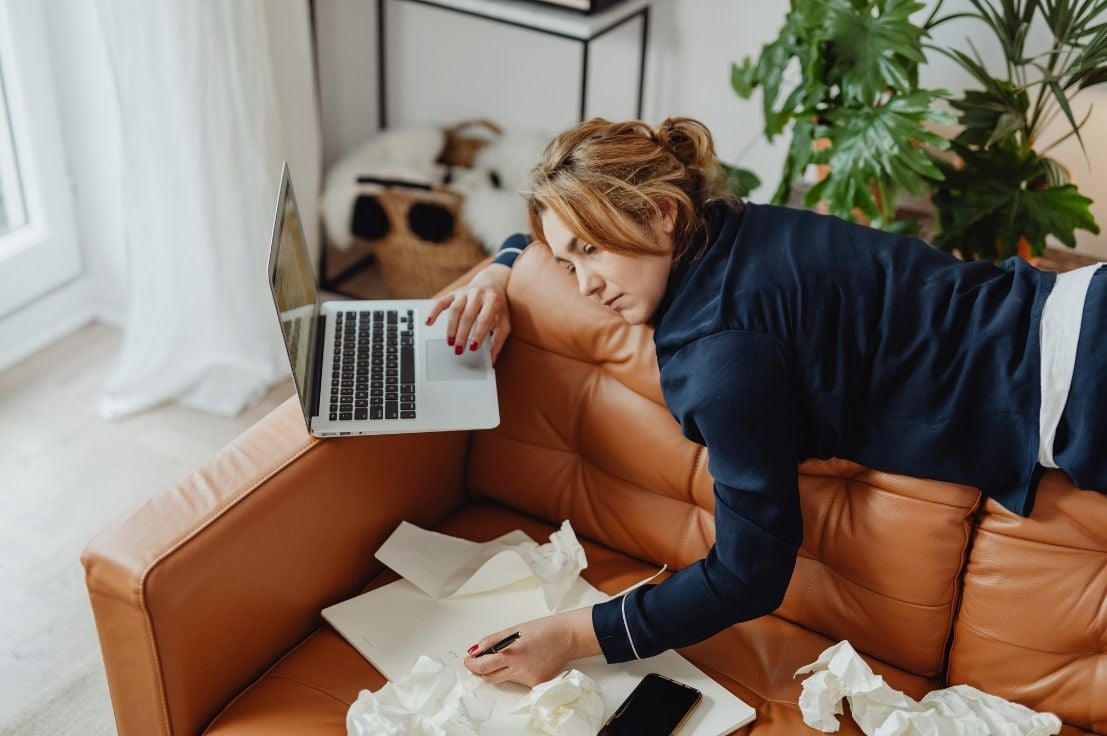 A woman under stress lays on a couch with a laptop and a lot of paper